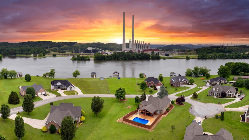 A residential area with houses, a swimming pool, and a large body of water with industrial structures in the background.