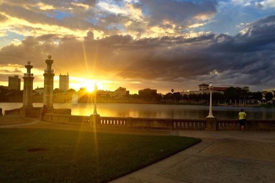 Un coucher de soleil sur une étendue d’eau, avec un parc et les contours d’une ville lointaine à l’horizon.