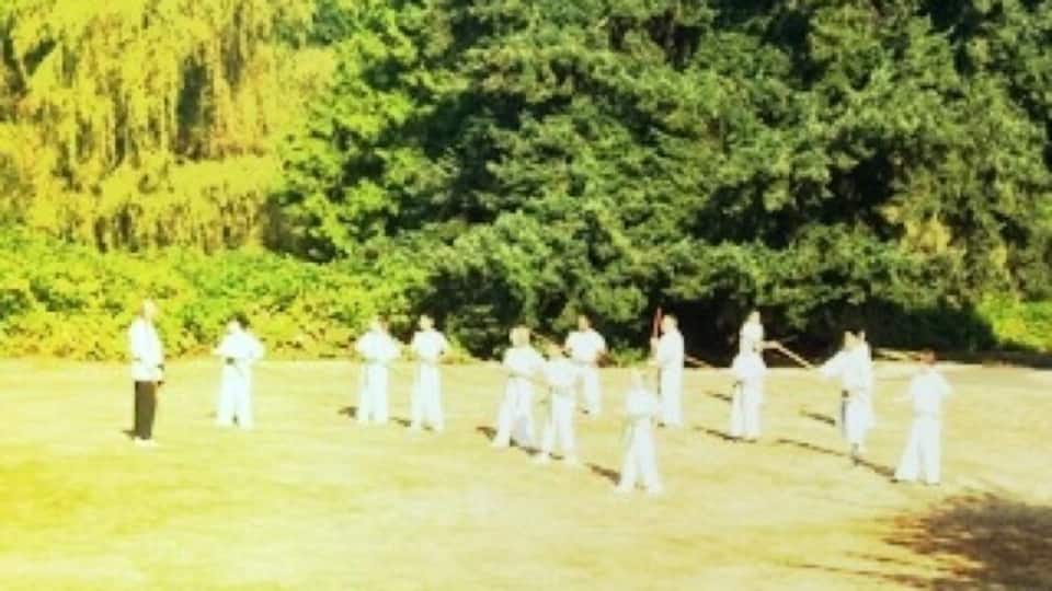 A group of people in white attire standing in a line on a grassy field with trees in the background.
