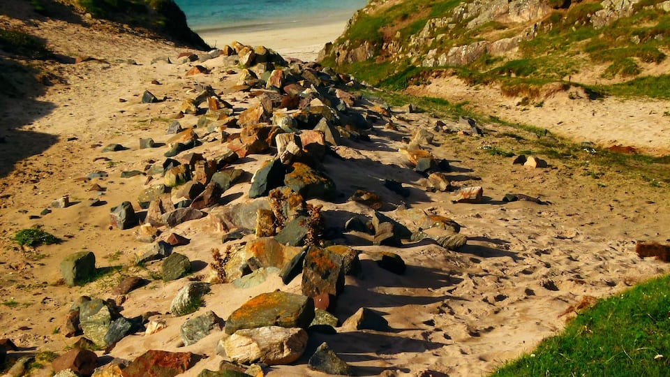 A rocky beach with a clear blue sea, grassy hills, and a few buildings in the distance.