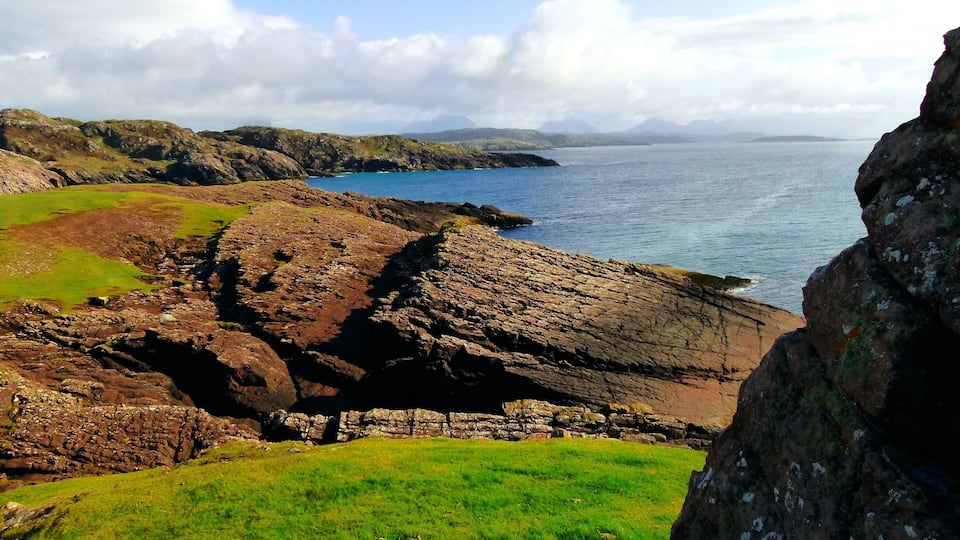 A coastal landscape with rocky terrain, grassy areas, and a view of the sea and distant mountains.