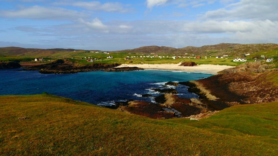 A coastal landscape with a beach, rocky shoreline, grassy hills, and scattered houses.