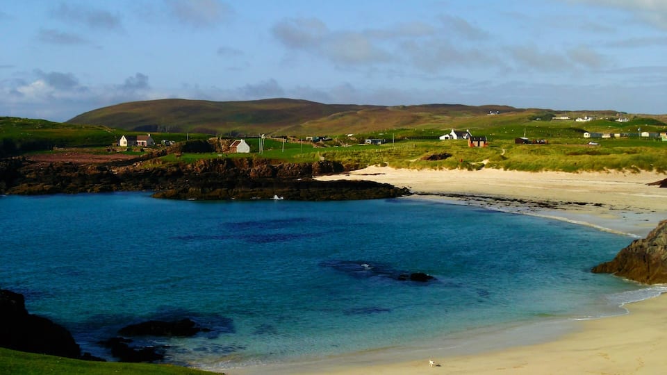 A coastal landscape with a sandy beach, calm blue waters, and a grassy hill with scattered houses.