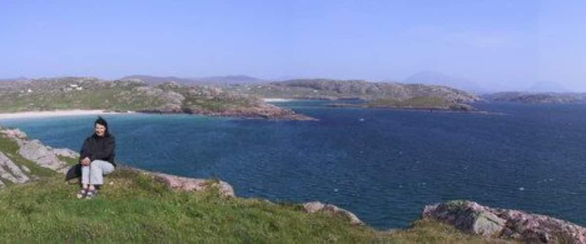 A person sitting on a grassy cliff overlooking a calm sea with distant hills.