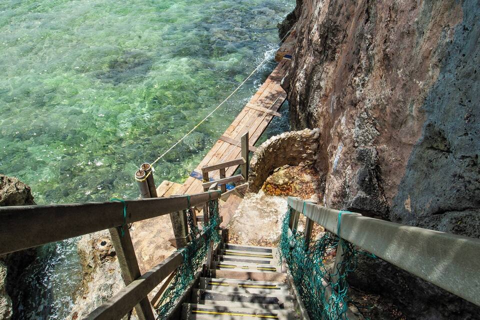 A staircase leading down to a clear body of water, surrounded by rocky cliffs and a wooden railing.