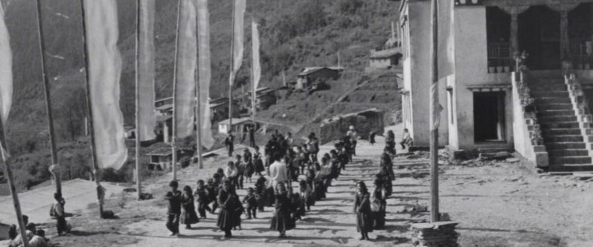 A group of people walking towards a building with a staircase, surrounded by flags and a mountainous landscape.