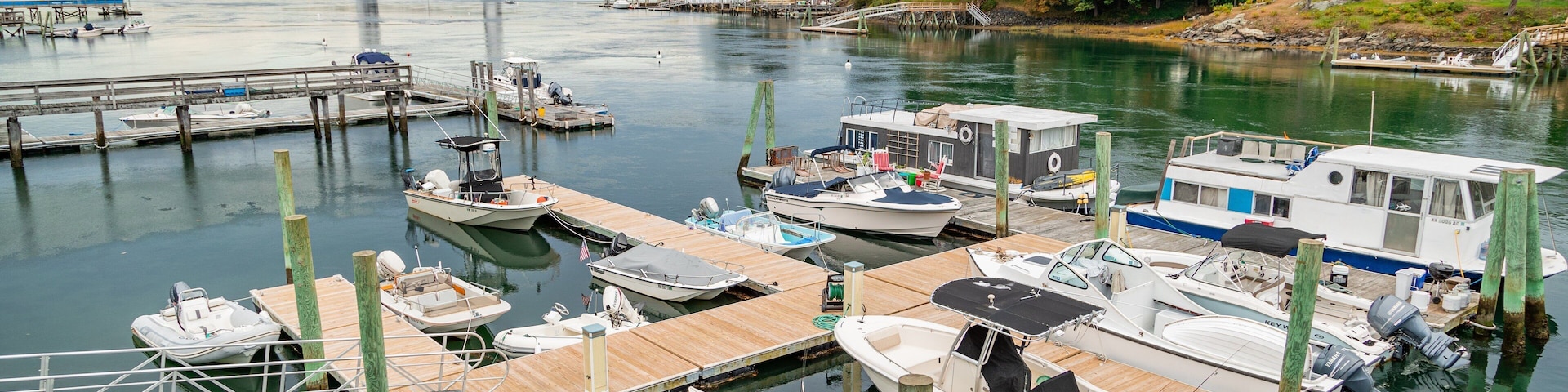 A marina with various boats docked at wooden piers, a bridge in the background, and a forested hillside.