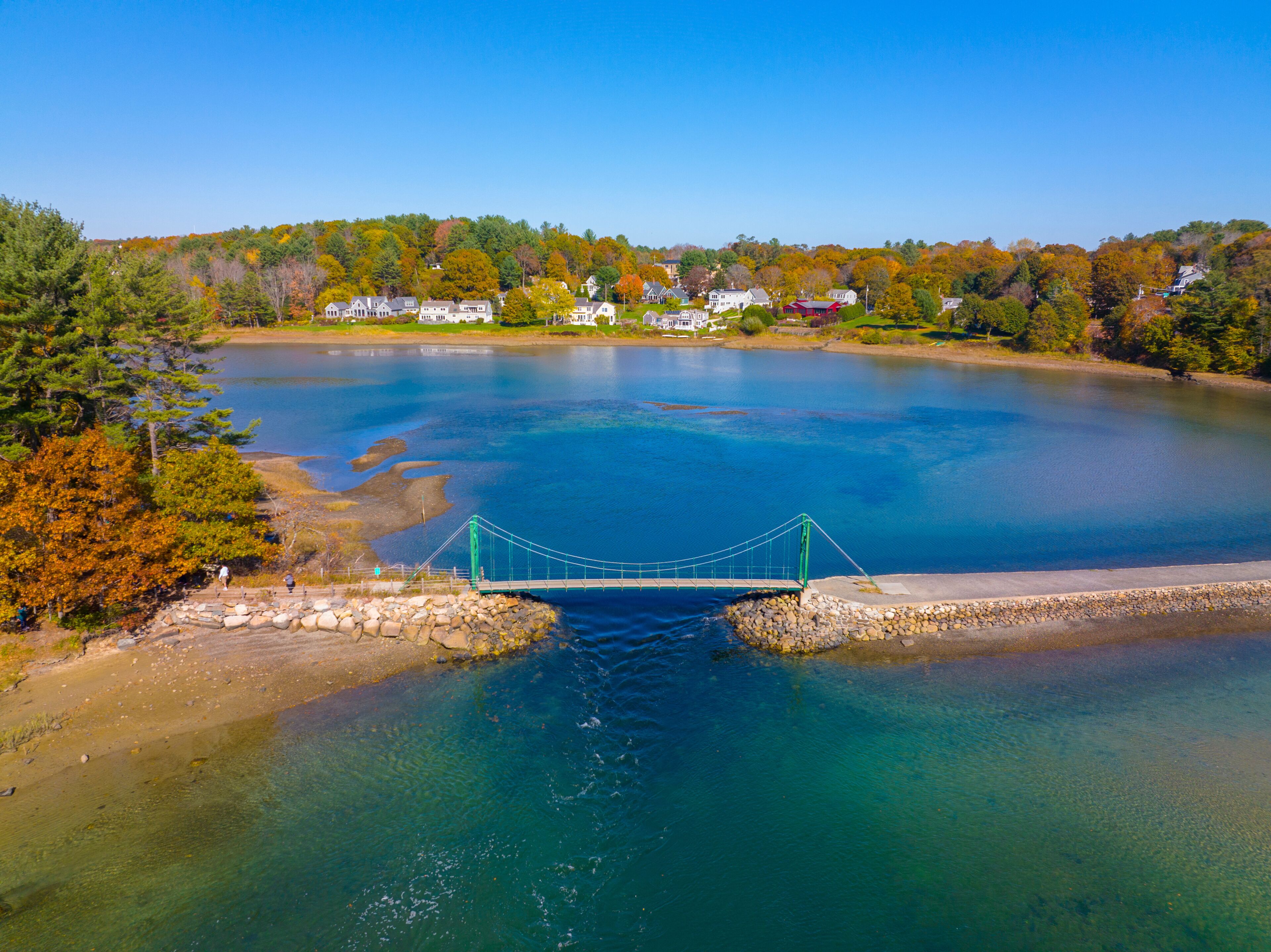 A suspension bridge over a calm lake with surrounding houses and autumn foliage.