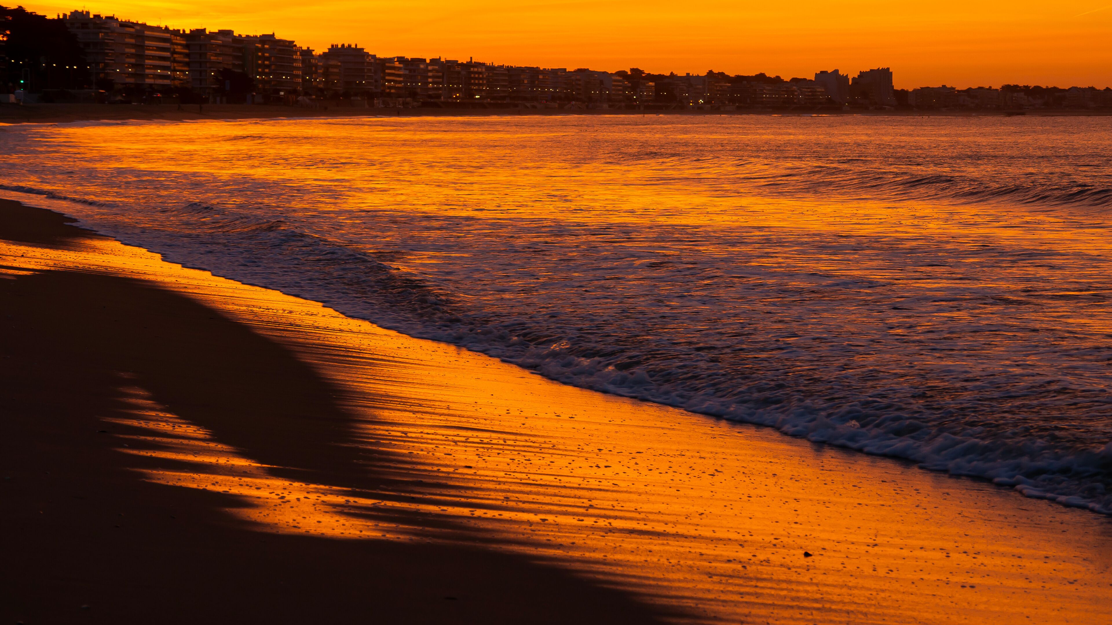A beach at sunset with waves, a sandy shore, and buildings in the background.