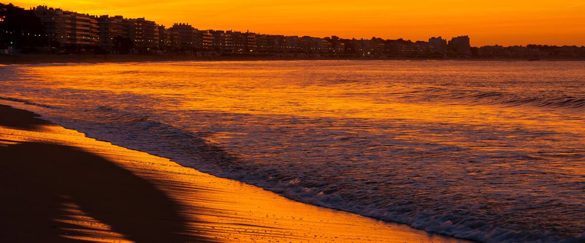 A beach at sunset with waves, a sandy shore, and buildings in the background.