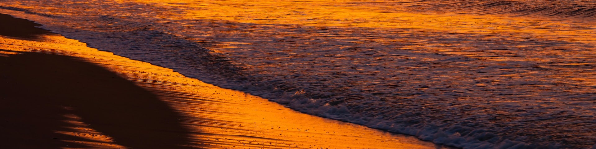 A beach at sunset with waves, a sandy shore, and buildings in the background.
