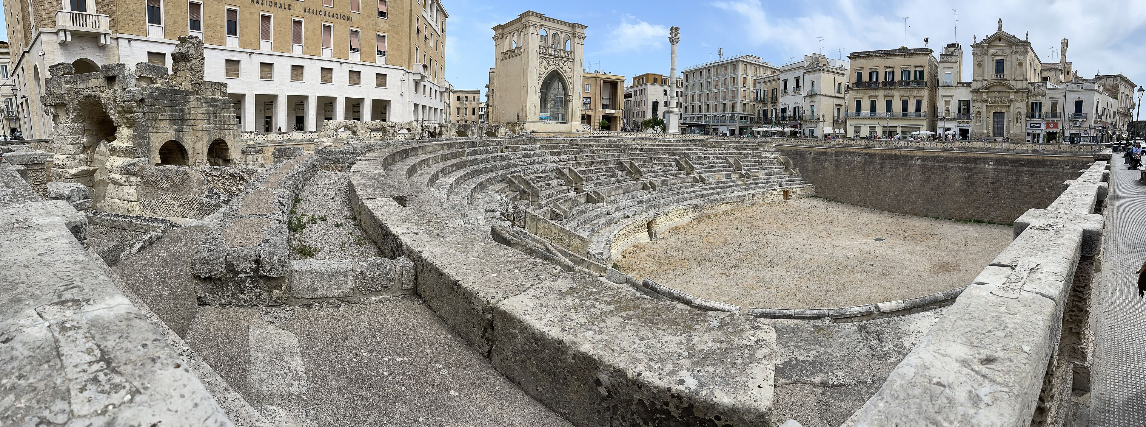 An ancient Roman amphitheater with stone steps and a central stage, surrounded by historic buildings.