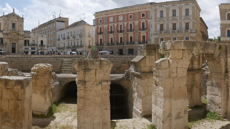 Ancient ruins with stone columns and arches, surrounded by historic buildings.