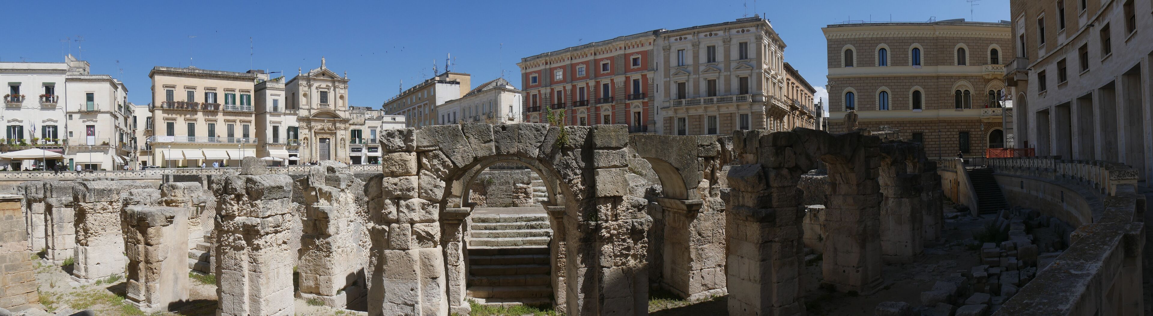 Ancient ruins with stone steps and arches, surrounded by historic buildings.