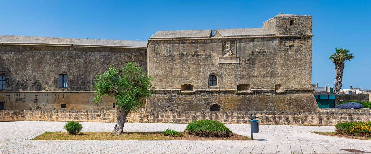 Une forteresse historique en pierre, dotée d’une tour centrale et de créneaux, entourée d’une cour pavée où se trouvent un palmier et de petits arbustes.