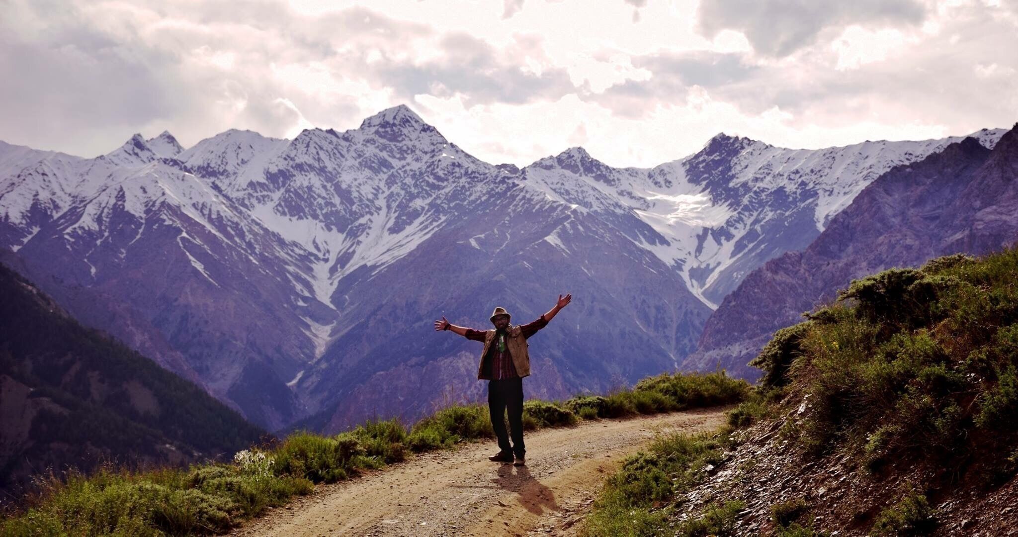 A person standing on a dirt path with arms outstretched, facing a mountain range with snow-capped peaks under a cloudy sky.