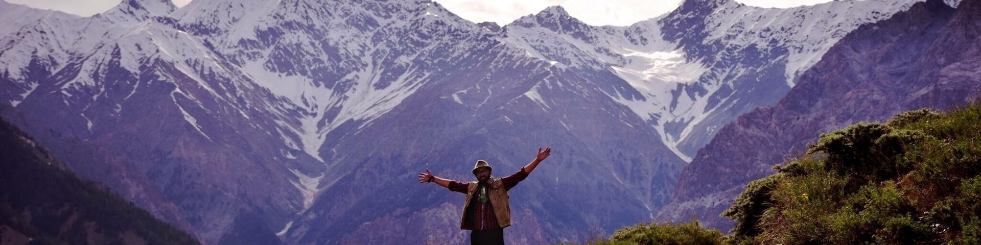 A person standing on a dirt path with arms outstretched, facing a mountain range with snow-capped peaks under a cloudy sky.