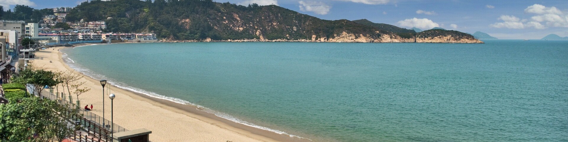 Una playa costera con aguas turquesas cristalinas, una orilla de arena y colinas y edificios al fondo.