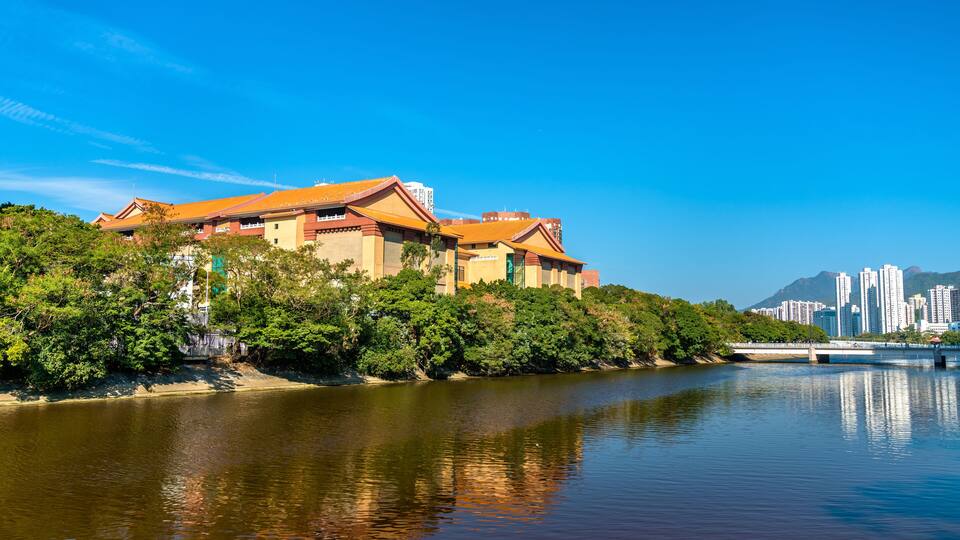 A riverside view with a traditional building, lush greenery, and a cityscape in the background.