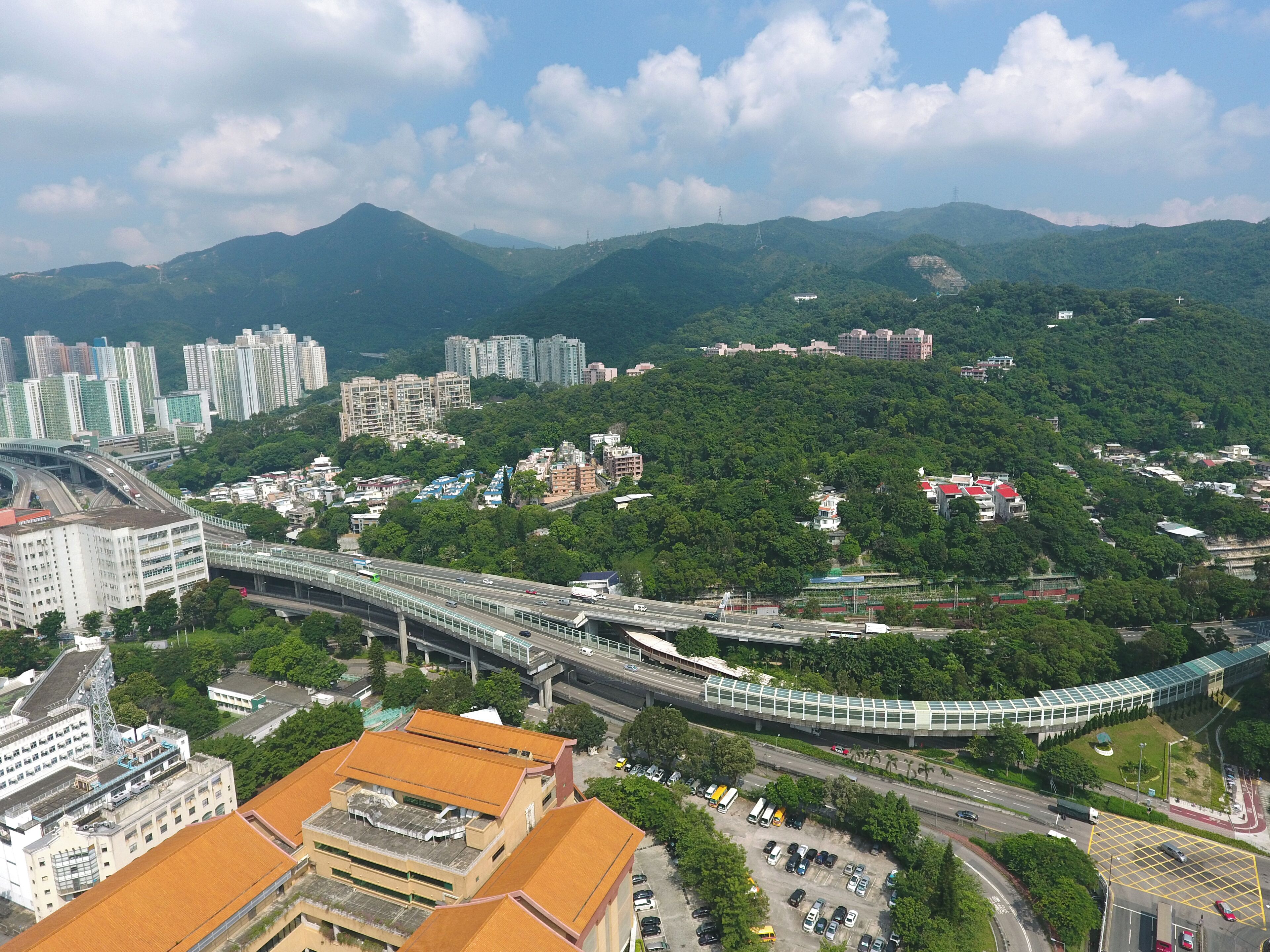 A cityscape with a modern elevated highway, dense urban buildings, and lush green hills in the background.