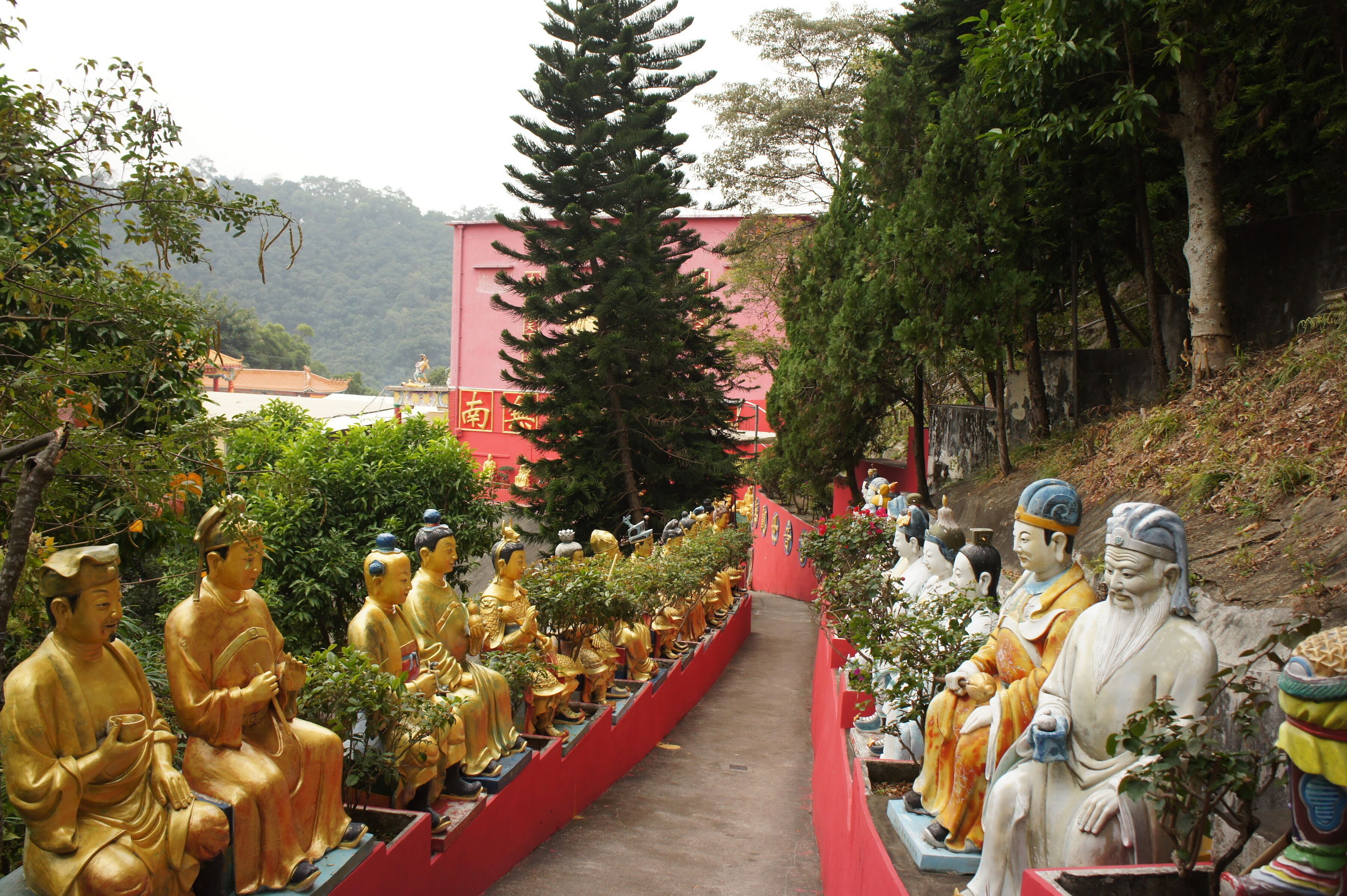 A row of golden Buddha statues along a red pathway, surrounded by trees and a building with Chinese characters.