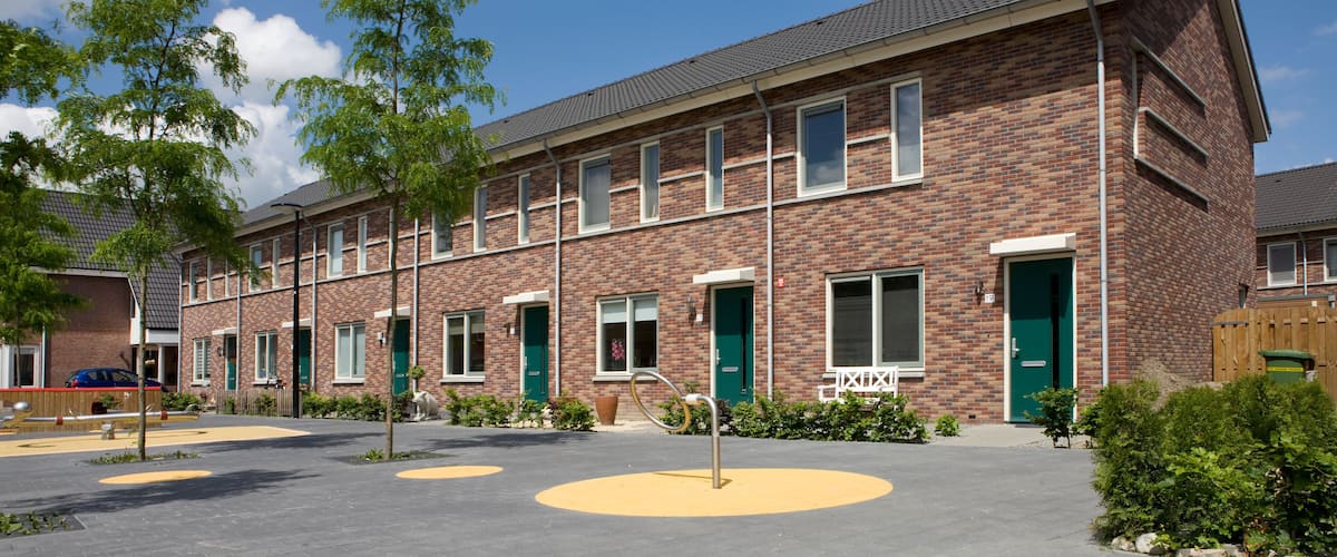 A row of brick buildings with green doors, a paved courtyard with yellow circles, and trees.