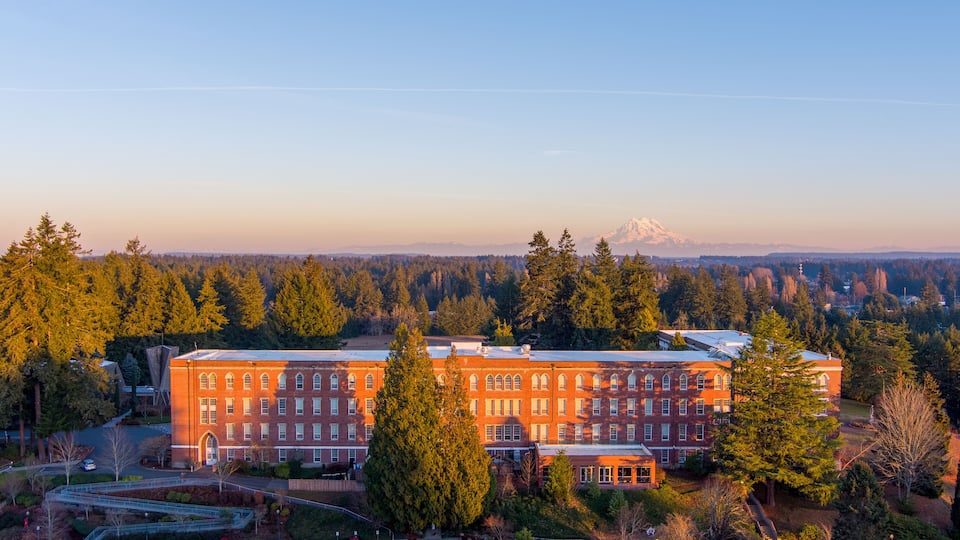 A large brick building surrounded by dense forest and a snow-capped mountain in the background.
