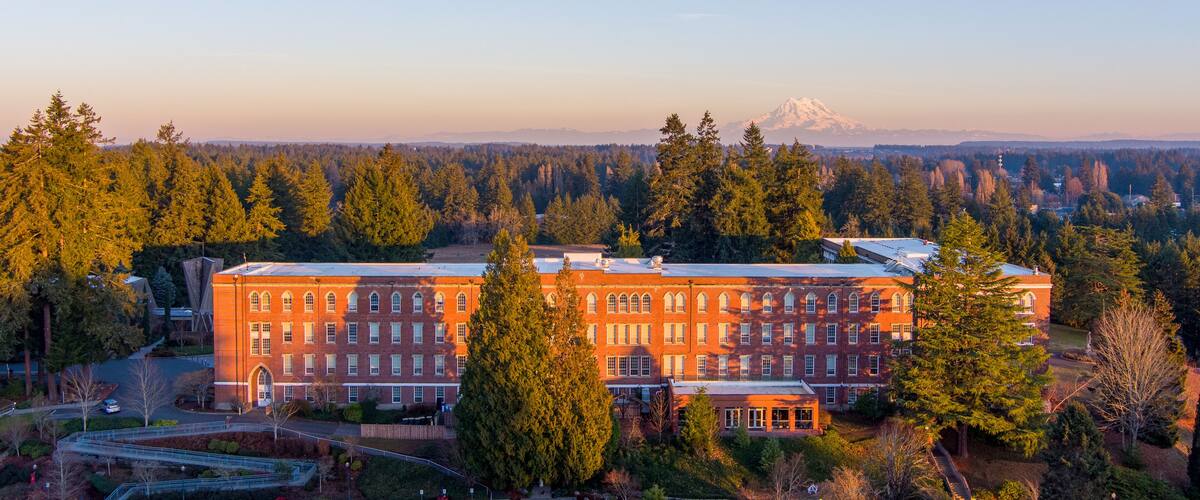 A large brick building surrounded by dense forest and a snow-capped mountain in the background.