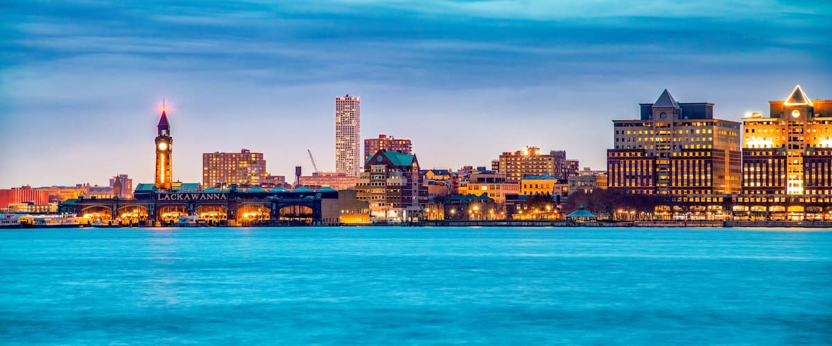 A waterfront cityscape with illuminated buildings, a prominent clock tower, and a clear blue sky.