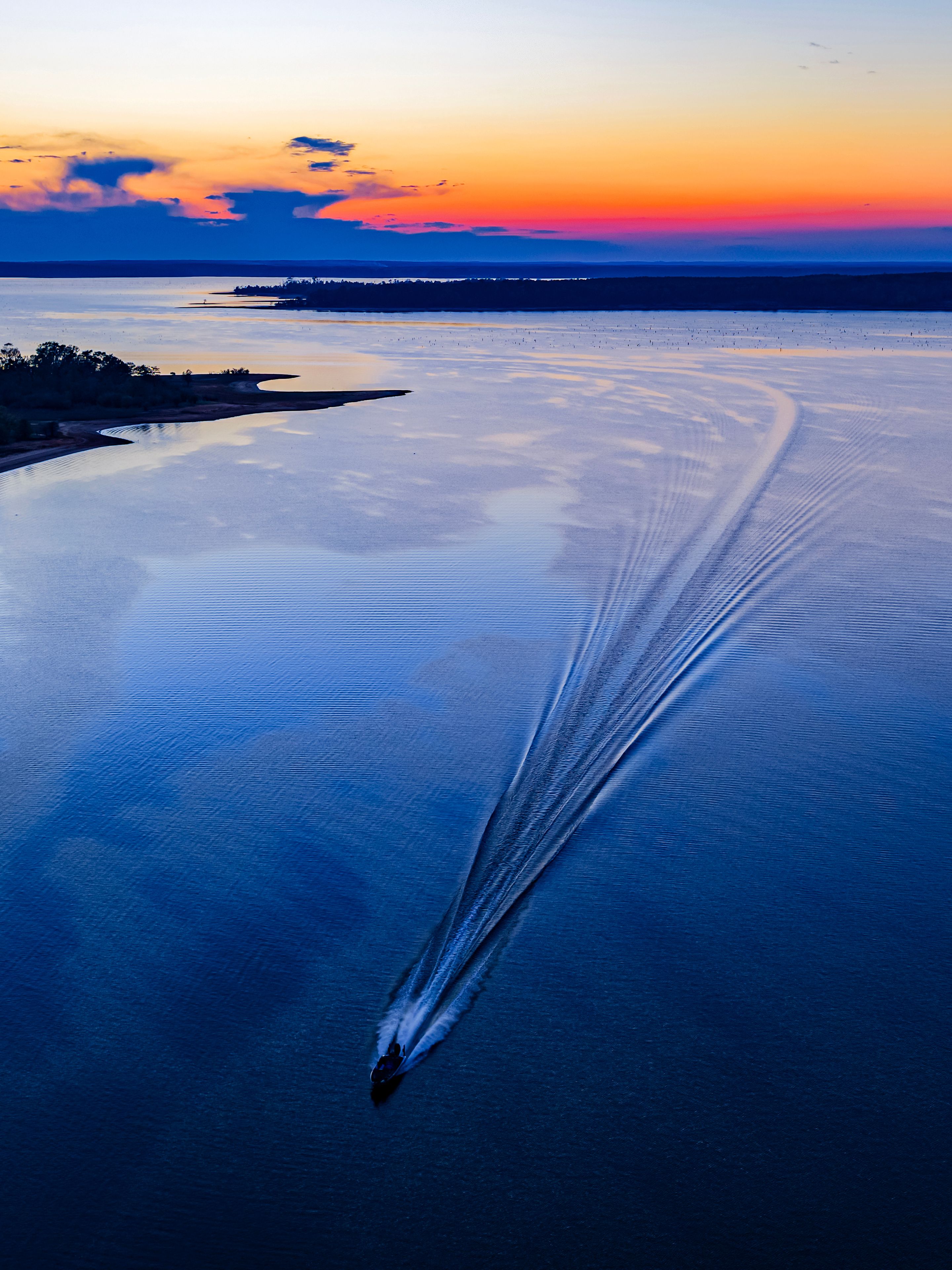 A body of water with a wake pattern, a distant shoreline, and a sunset sky.