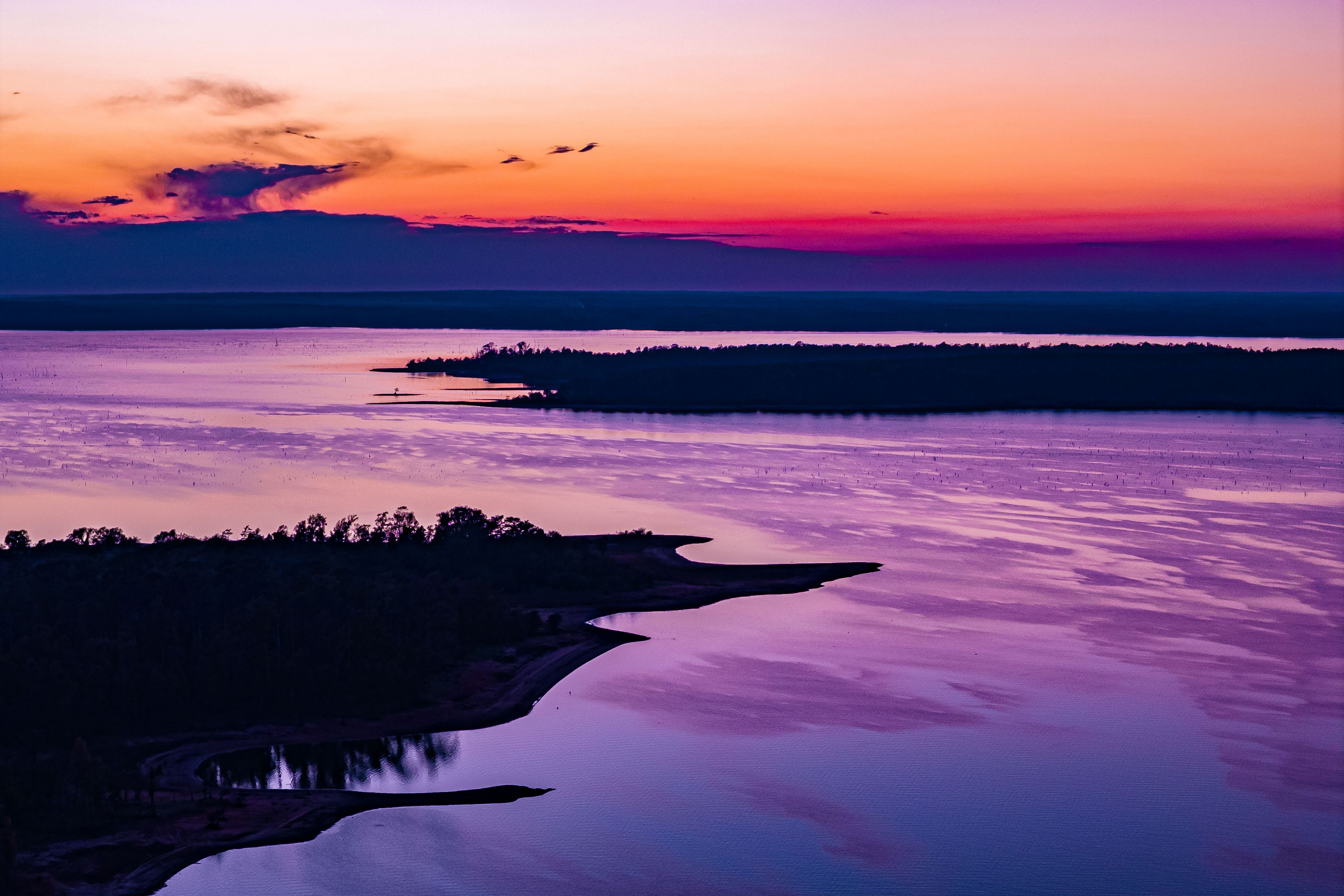 Ein Sonnenuntergang über einem stillen See mit bewaldeter Uferlinie und entfernten Landformen.