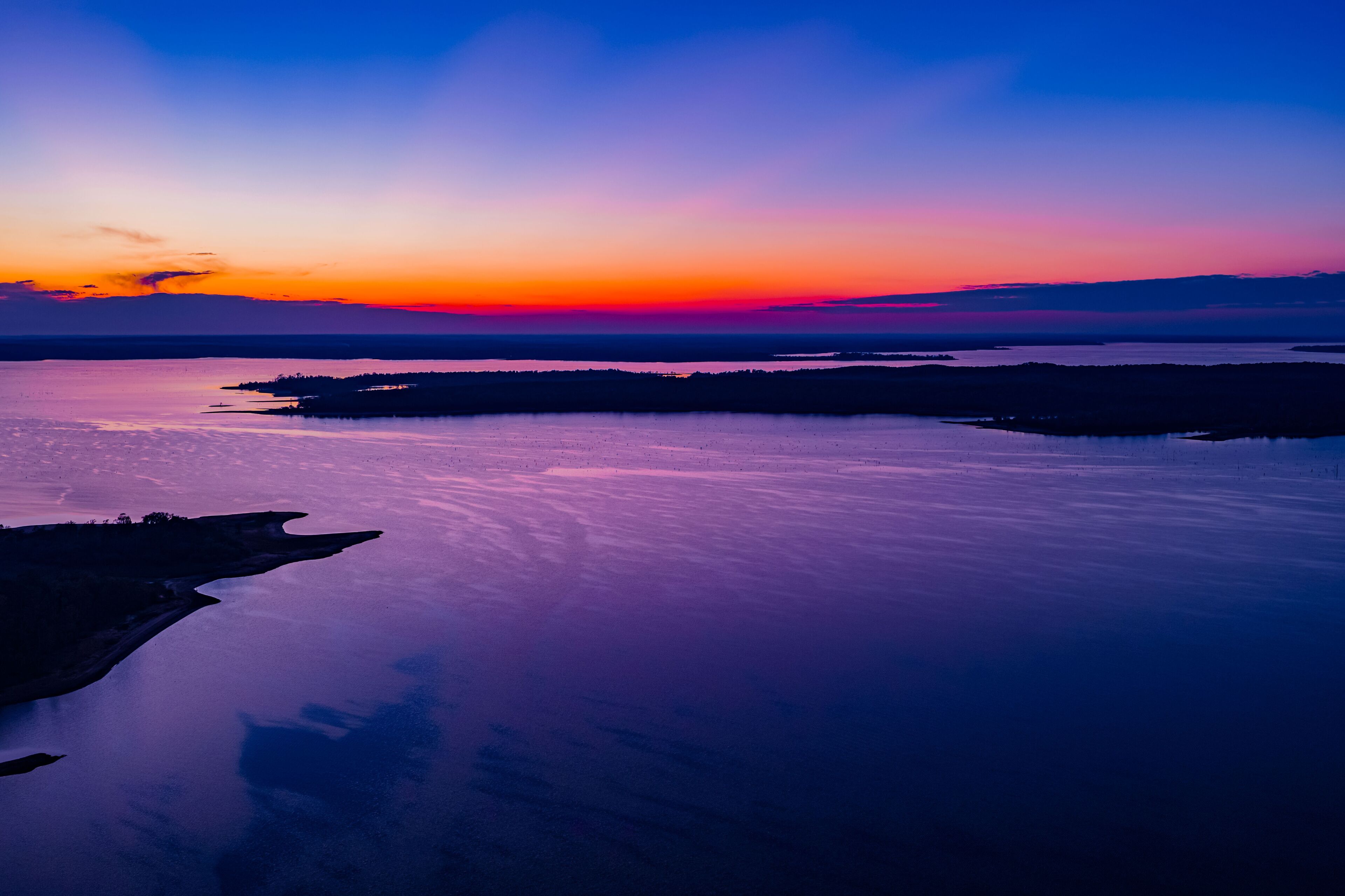 A sunset over a calm lake with a silhouette of a peninsula and distant land.