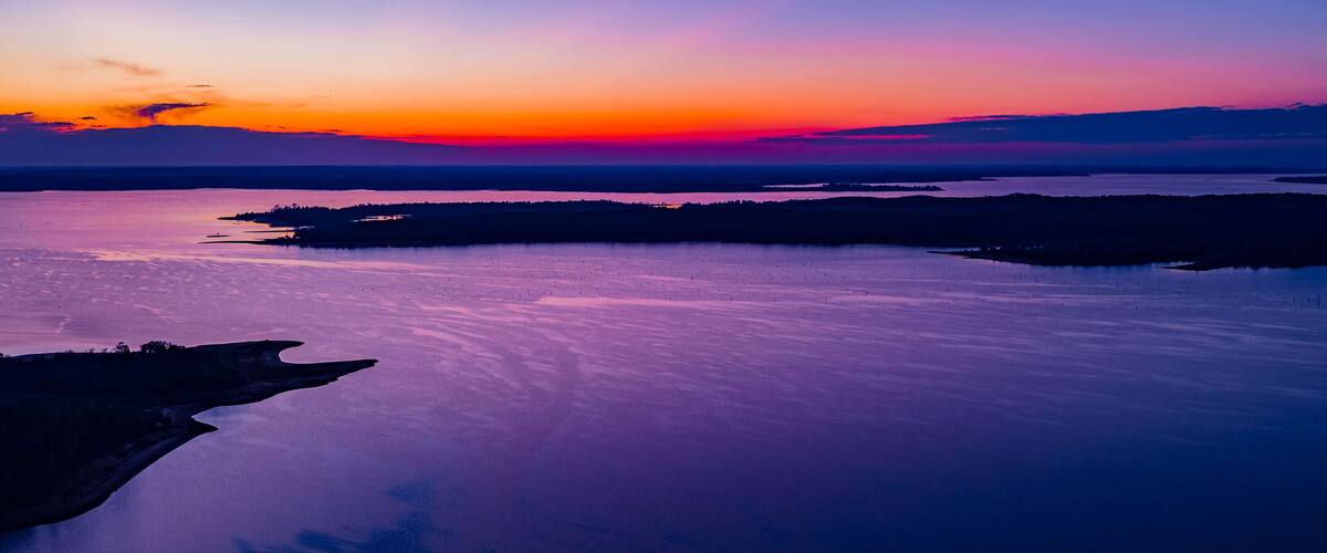 A sunset over a calm lake with a silhouette of a peninsula and distant land.