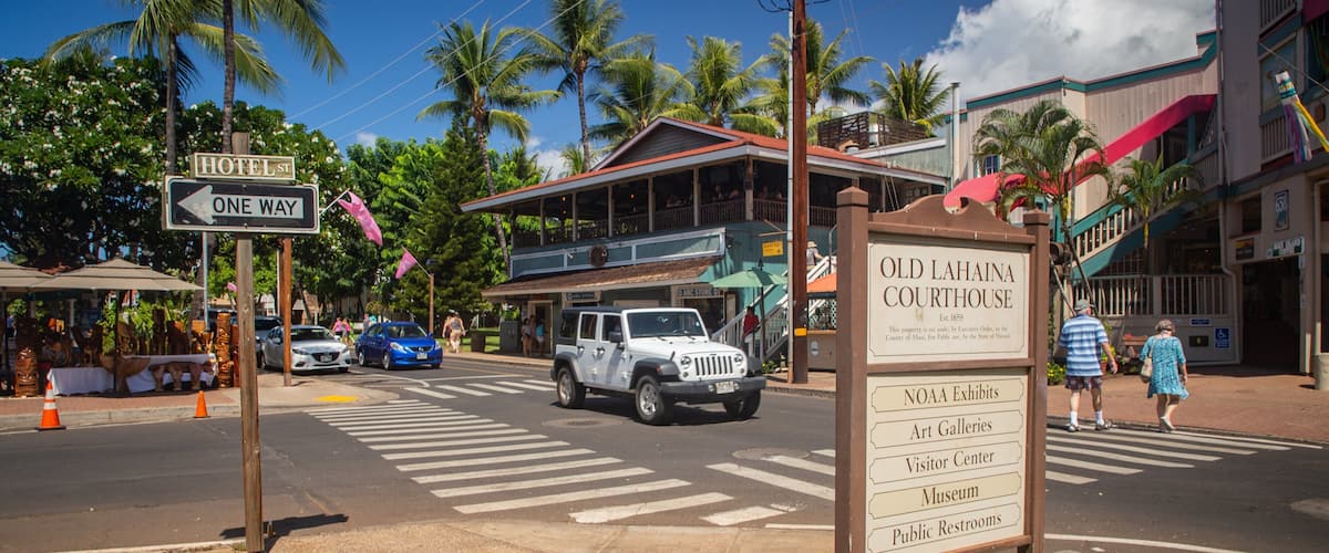 Een straat met een bord voor het Old Lahaina Courthouse, een blauwe auto en palmbomen.