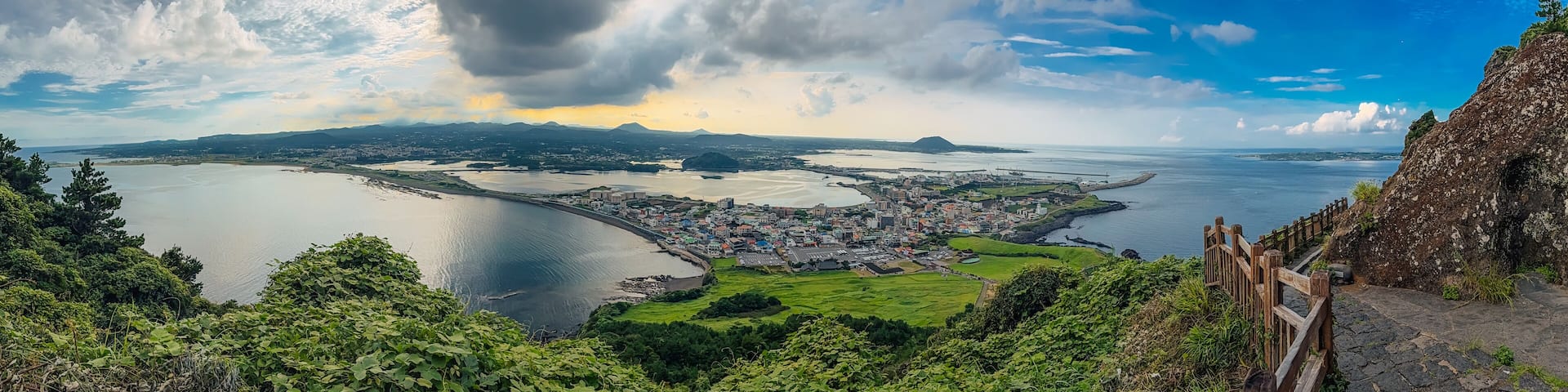 Ultra-wide panoramic coastal scenery on Jeju Island, South Korea, showing a town, curved shoreline, green hills, and a dramatic sky, framed by a cliffside path and railing.
