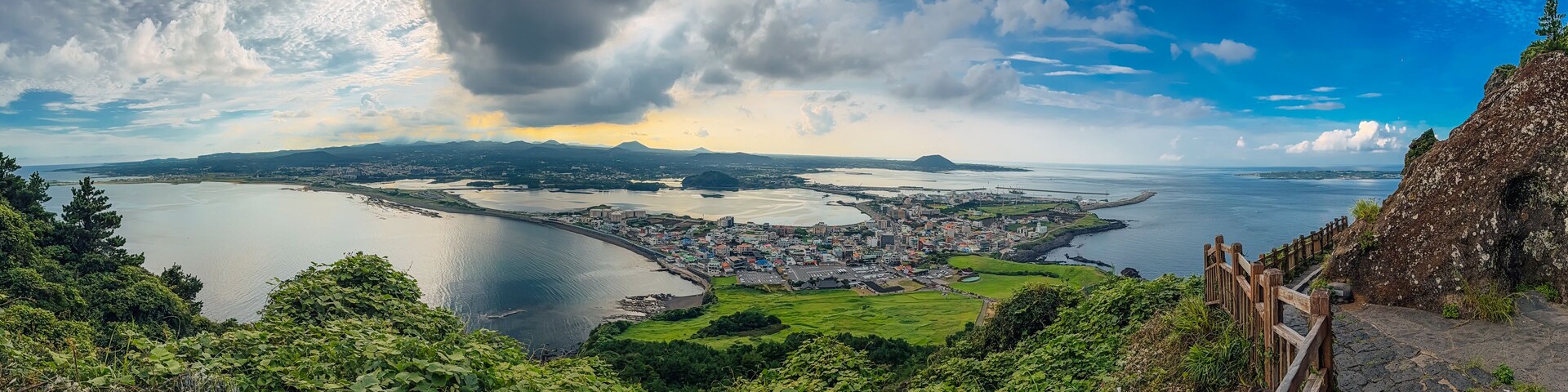 Ultra-wide panoramic coastal scenery on Jeju Island, South Korea, showing a town, curved shoreline, green hills, and a dramatic sky, framed by a cliffside path and railing.