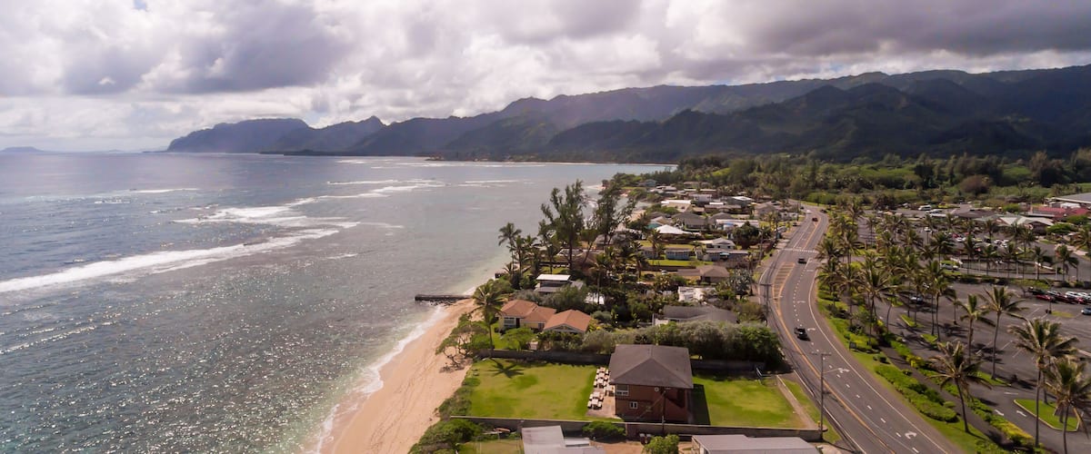 A coastal town with a beach, houses, palm trees, and mountains in the background.