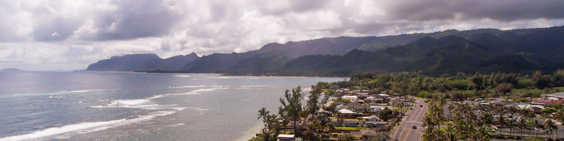 A coastal town with a beach, houses, palm trees, and mountains in the background.