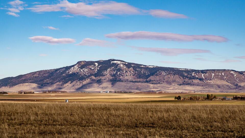 A mountain range with snow-capped peaks, a clear blue sky, and a flat agricultural field in the foreground.