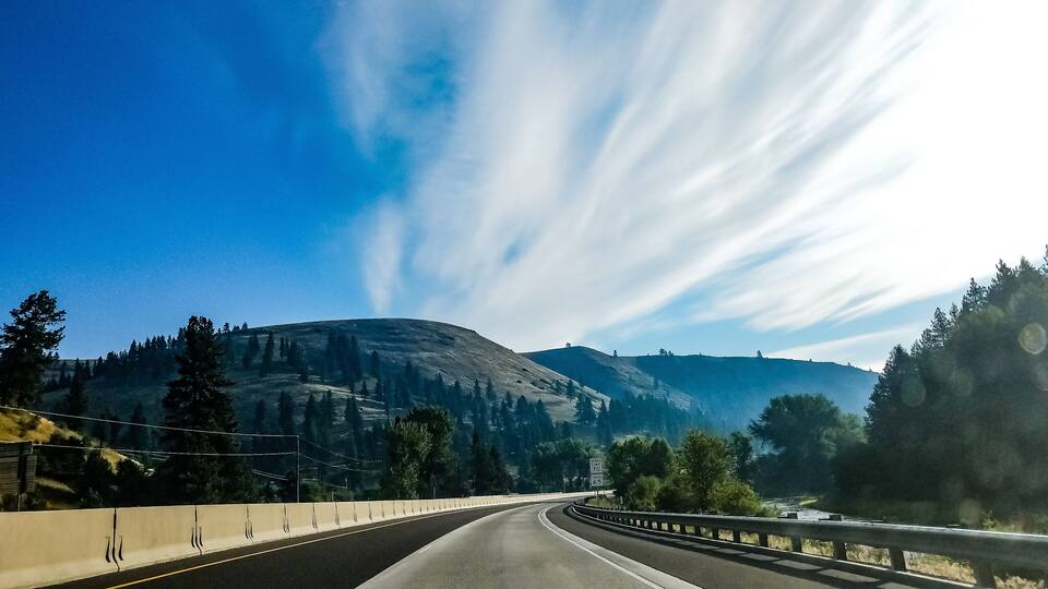 A winding highway with a guardrail, surrounded by mountains and trees under a clear blue sky.