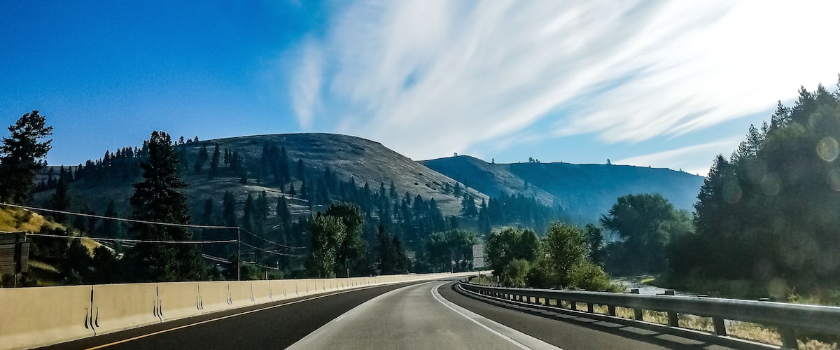 A winding highway with a guardrail, surrounded by mountains and trees under a clear blue sky.