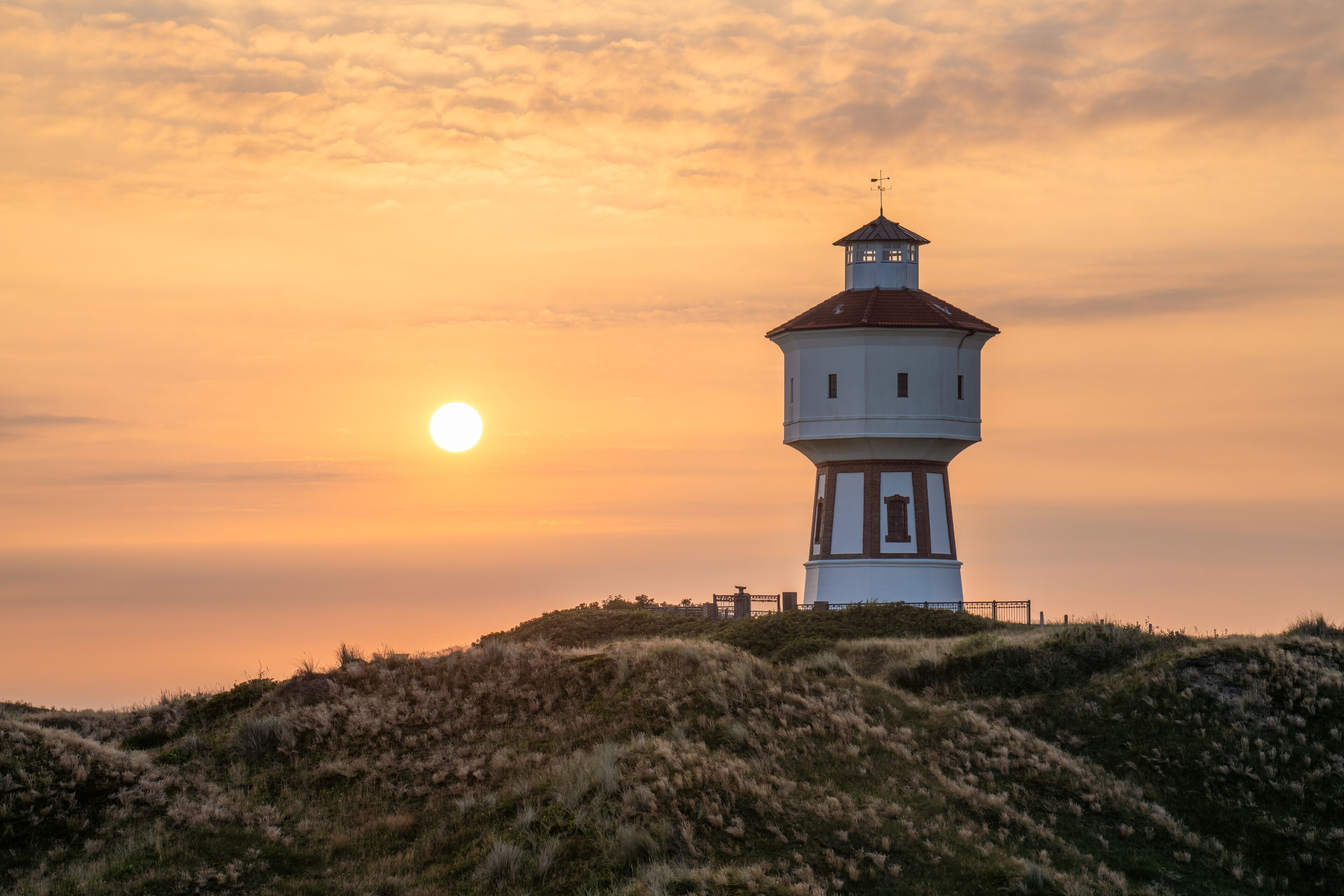 A lighthouse on a grassy hill with the sun setting in the background.