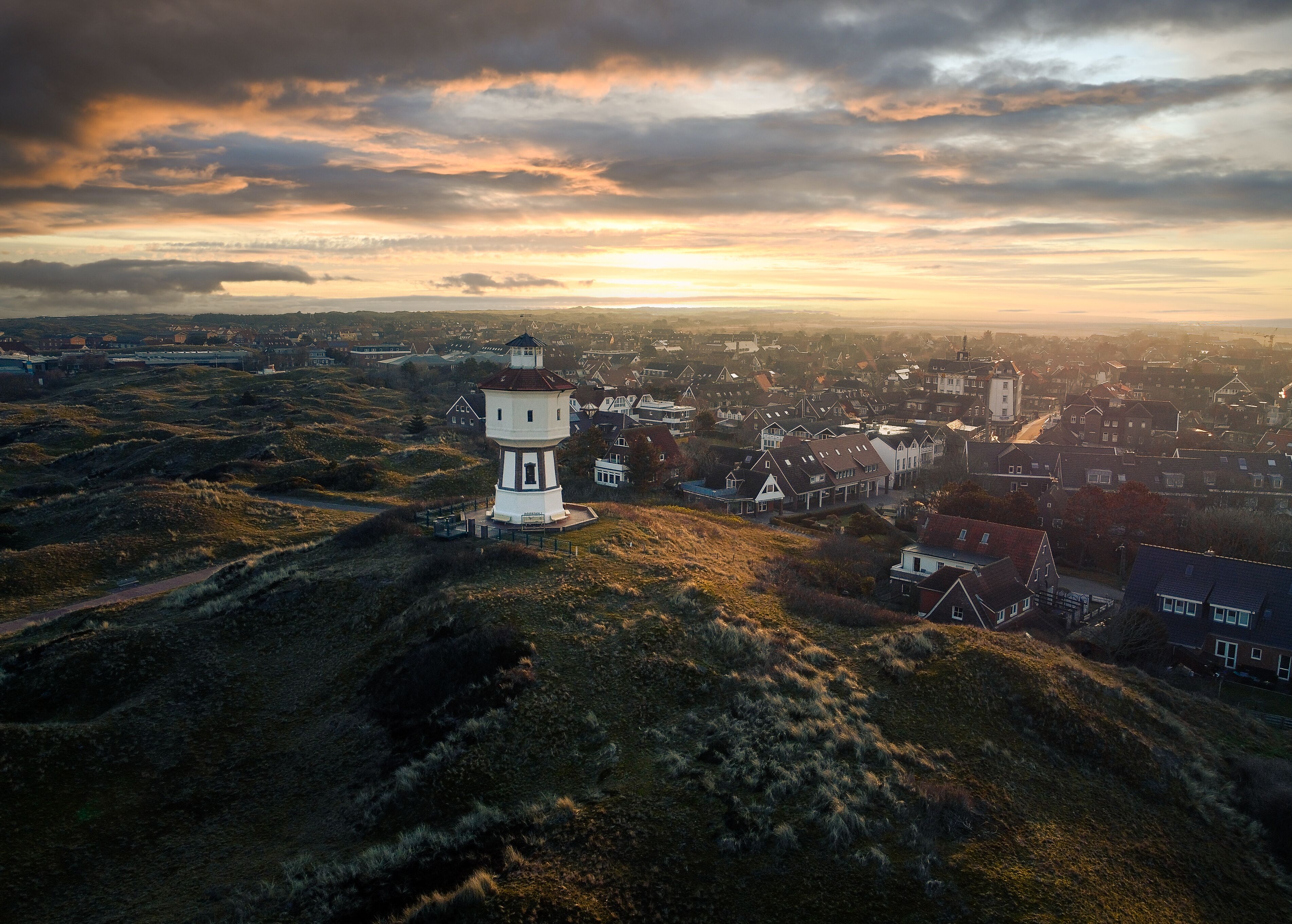 A lighthouse on a hill overlooking a coastal town with houses and a sunset sky.