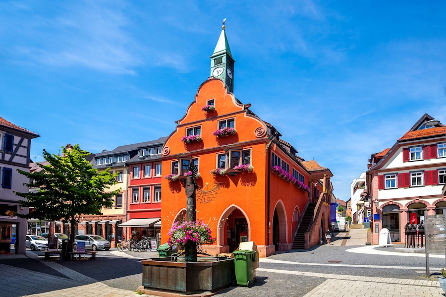 Un bâtiment d’un orange éclatant, surmonté d’une flèche verte, entouré d’une architecture européenne traditionnelle.