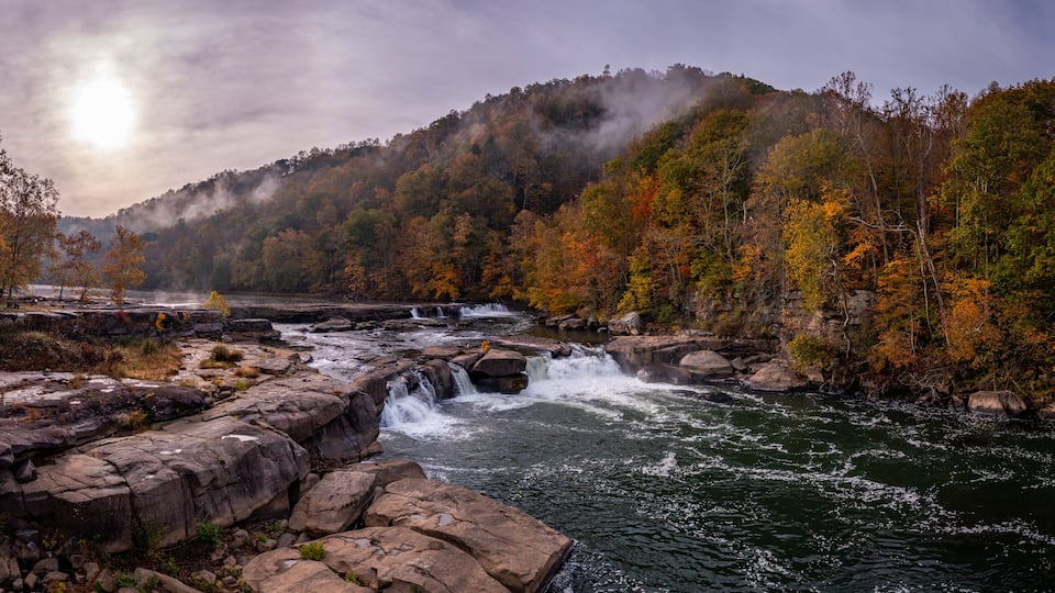 A river with a waterfall, surrounded by autumn-colored trees and a misty mountain in the background.