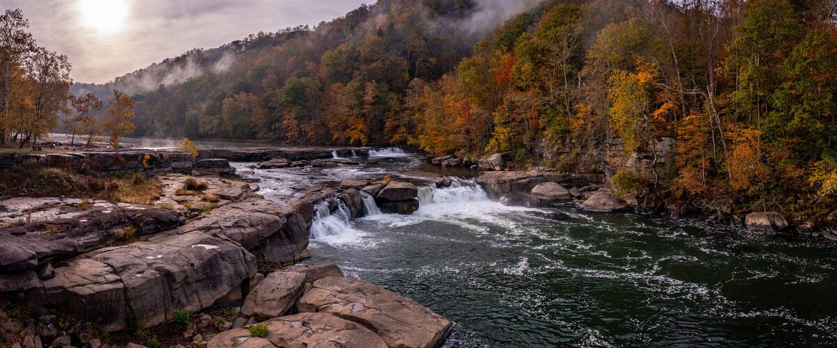 A river with a waterfall, surrounded by autumn-colored trees and a misty mountain in the background.