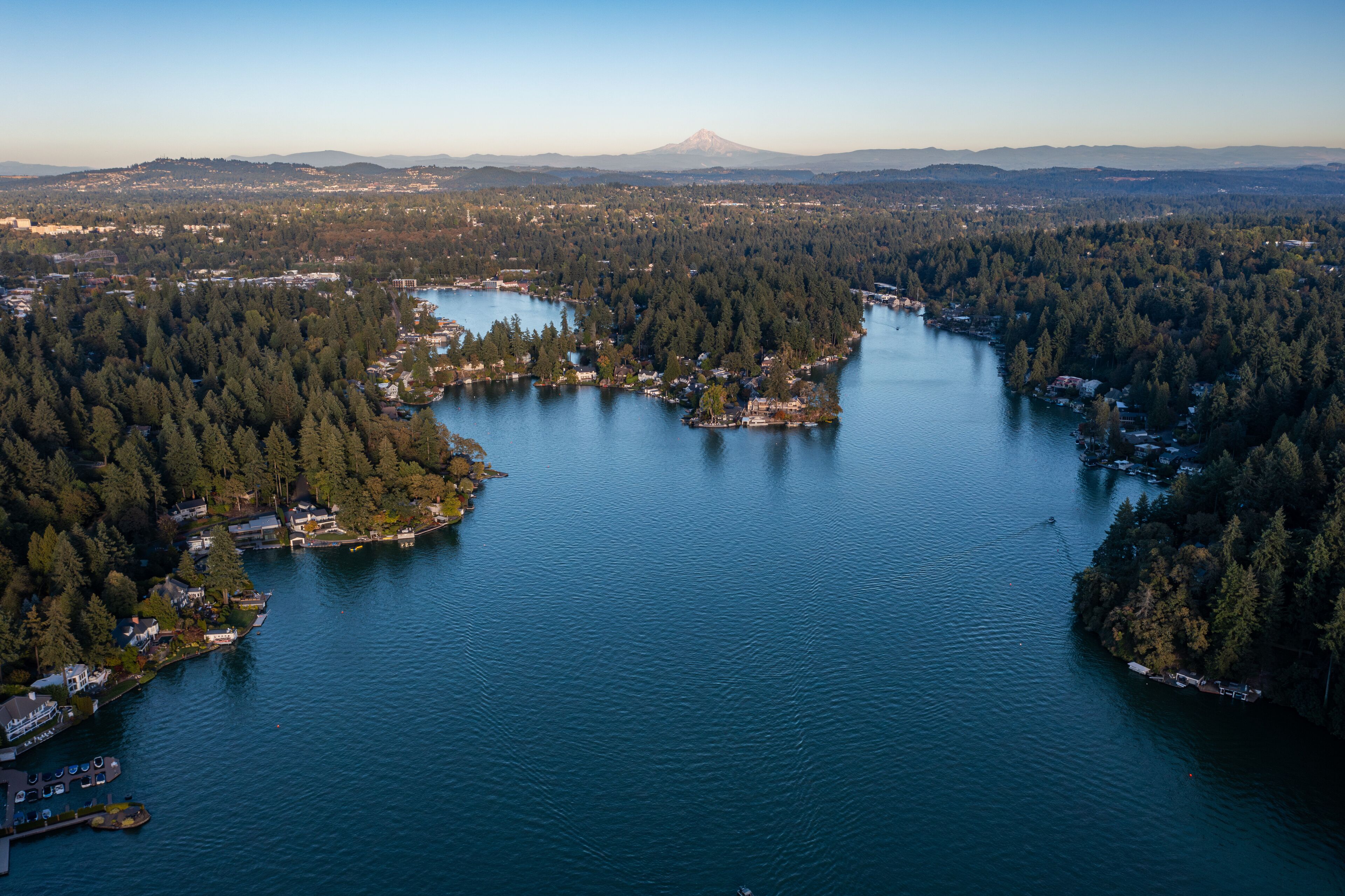 Aerial drone picture of Lake Oswego, Oregon and Mount Hood in backgrond, showing calm blue water surrounded by dense forest and residential houses, real estate, on a clear sunny evening at sunset.  