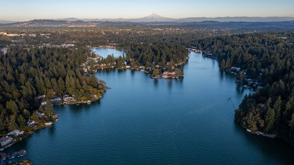 Aerial drone picture of Lake Oswego, Oregon and Mount Hood in backgrond, showing calm blue water surrounded by dense forest and residential houses, real estate, on a clear sunny evening at sunset.