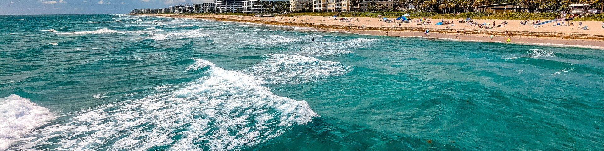 Escena costera con aguas turquesas, una playa de arena y edificios frente al mar.