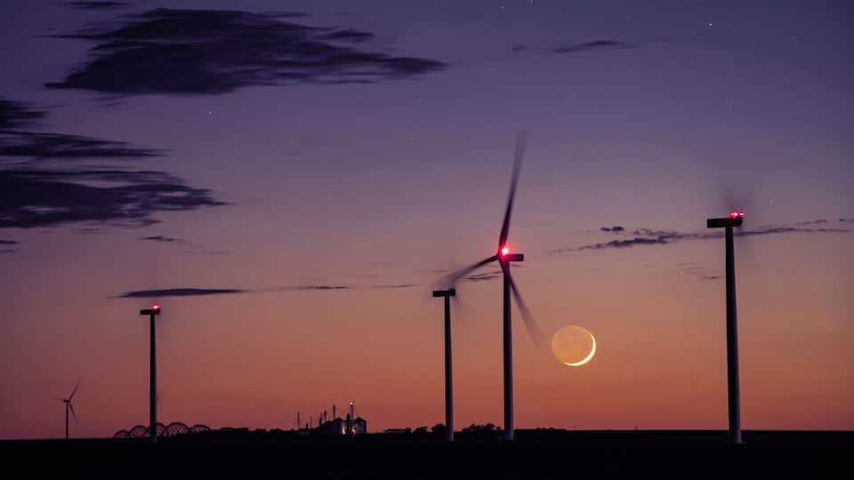Wind turbines at sunset with a crescent moon in the sky.